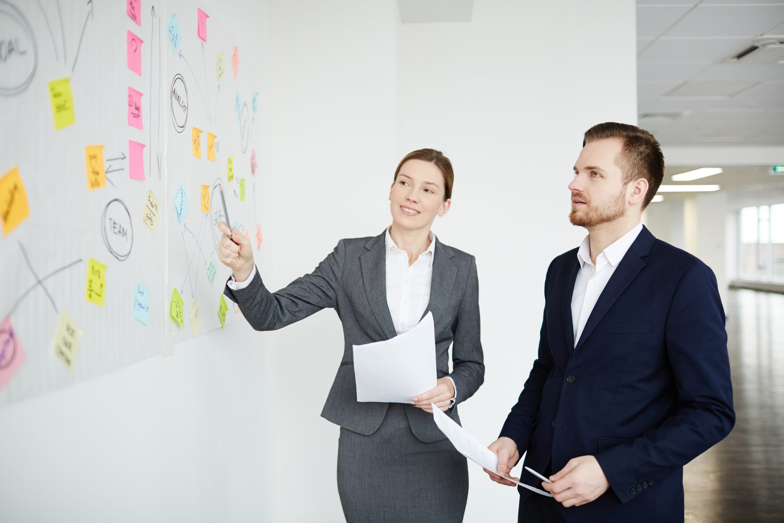 Businesswoman pointing at one of notepapers on whiteboard while discussing working points with colleague
